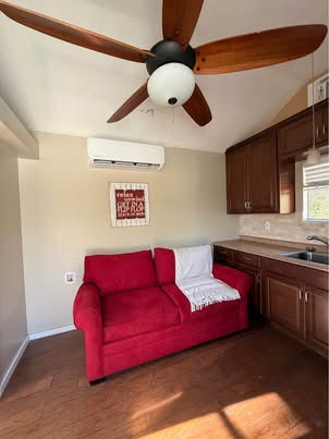 Small cottage living space with red sofa, kitchenette cabinets, ceiling fan, and sunlight.